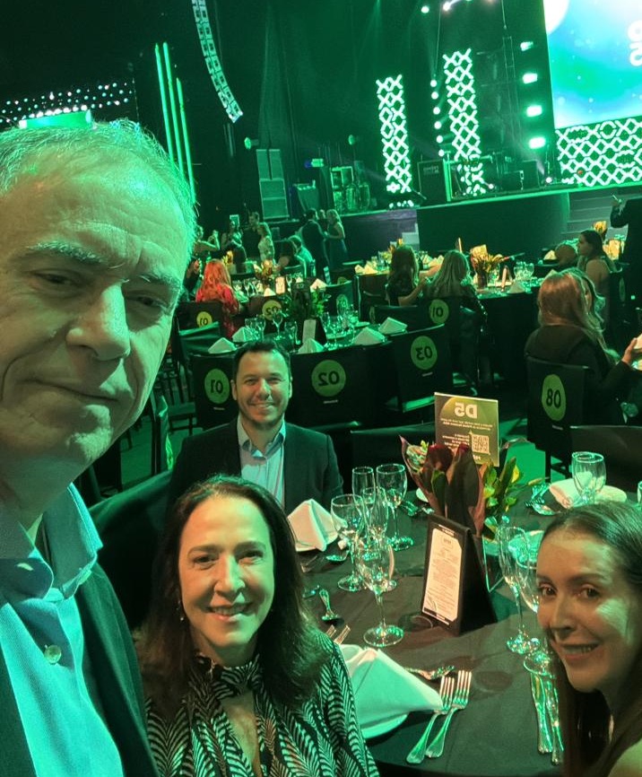 Quatro pessoas reunidas em uma mesa em um evento de gala iluminado em tons de verde, com palco iluminado ao fundo e mesas elegantemente preparadas